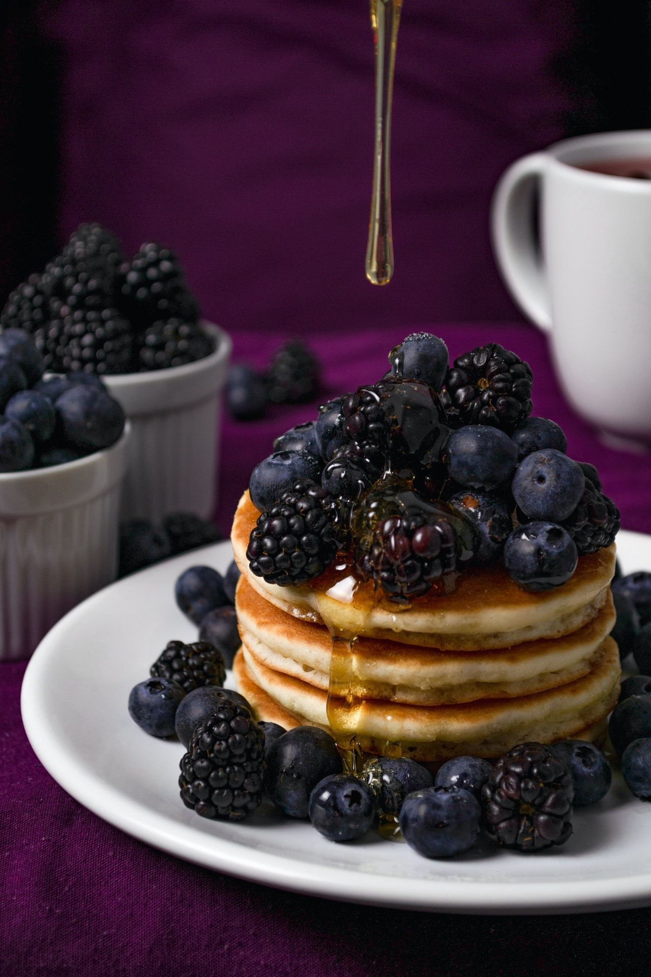 Stack of pancakes topped with blueberries and blackberries with syrup being poured on them with a purple background